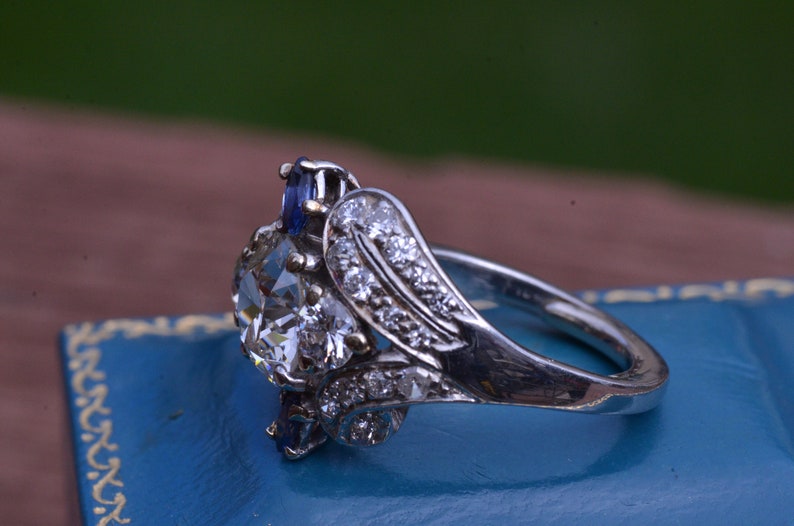 May include: A silver ring featuring a large, clear, round diamond surrounded by smaller diamonds and two blue sapphires.