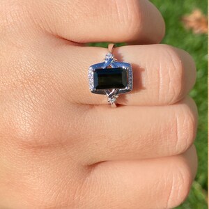 K&ouml;nnte beinhalten: Ein silberner Ring mit einem schwarzen Smaragdschliff-Edelstein, umgeben von kleineren klaren Edelsteinen.
