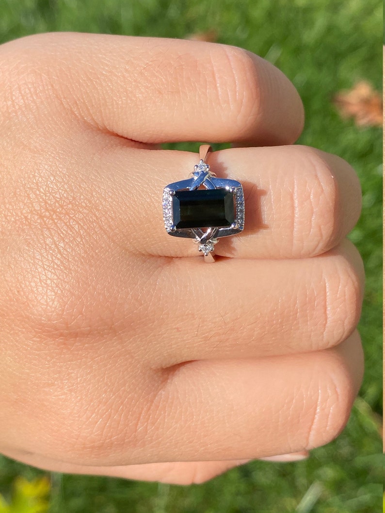 K&ouml;nnte beinhalten: Ein silberner Ring mit einem schwarzen Smaragdschliff-Edelstein, umgeben von kleineren klaren Edelsteinen.