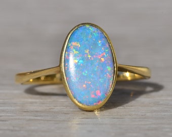 Puede incluir: Anillo de oro con una piedra de ópalo ovalada. El ópalo tiene un color base azul claro con destellos rojos, amarillos y verdes. El anillo tiene una banda de oro pulido.