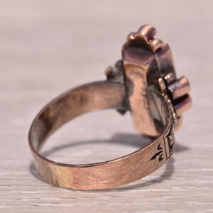 Pu&ograve; includere: Un anello vintage color rame con una montatura rettangolare ornata. La fascia dell'anello presenta dettagli incisi e la montatura mostra una pietra centrale con accenti circostanti. L'anello &egrave; esposto su una superficie chiara.