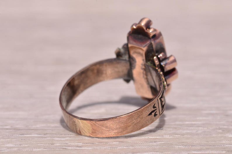 Pu&ograve; includere: Un anello vintage color rame con una montatura rettangolare ornata. La fascia dell'anello presenta dettagli incisi e la montatura mostra una pietra centrale con accenti circostanti. L'anello &egrave; esposto su una superficie chiara.