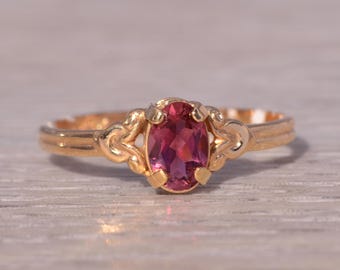 Puede incluir: Un anillo de oro con una piedra preciosa ovalada de color rosa intenso. El anillo tiene un diseño ornamentado alrededor de la piedra preciosa y una banda simple. El anillo está sobre una superficie clara y texturizada. Joyería.