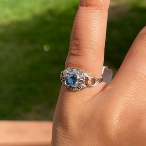 K&ouml;nnte beinhalten: Ein silberner Ring mit einem blauen Edelstein, der von kleineren, klaren Edelsteinen umgeben ist.