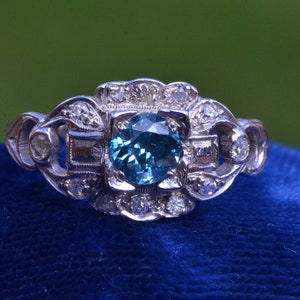 K&ouml;nnte beinhalten: Ein silberner Ring mit einem blauen Edelstein, der von kleineren Diamanten umgeben ist. Der Ring hat ein zartes, filigranes Design.