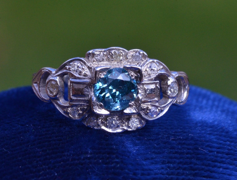 K&ouml;nnte beinhalten: Ein silberner Ring mit einem blauen Edelstein, der von kleineren Diamanten umgeben ist. Der Ring hat ein zartes, filigranes Design.