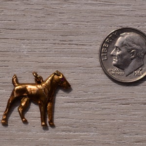 May include: A gold-colored metal charm of a wirehaired fox terrier dog. The charm is about the size of a dime.