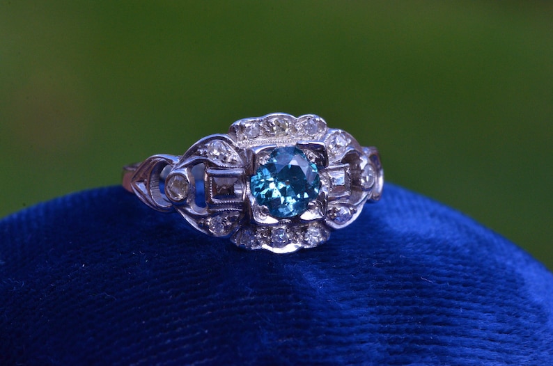 K&ouml;nnte beinhalten: Ein silberner Ring mit einem blauen Edelstein, der von kleineren Diamanten umgeben ist. Der Ring hat ein zartes, filigranes Design.