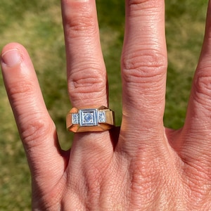 以下が含まれることがあります： 正方形の中央の石と、両側に2つの小さな石が付いた金の指輪。指輪は人の指にはめられています。