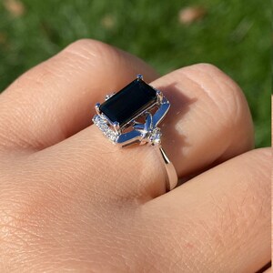 K&ouml;nnte beinhalten: Ein silberner Ring mit einem gro&szlig;en, dunkelgr&uuml;nen, rechteckigen schwarzen Edelstein, der von kleineren, klaren Edelsteinen umgeben ist. Der Ring wird an einem Finger getragen.