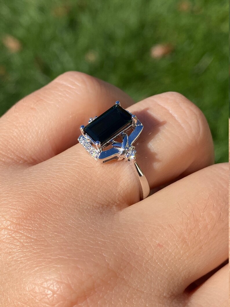 K&ouml;nnte beinhalten: Ein silberner Ring mit einem gro&szlig;en, dunkelgr&uuml;nen, rechteckigen schwarzen Edelstein, der von kleineren, klaren Edelsteinen umgeben ist. Der Ring wird an einem Finger getragen.