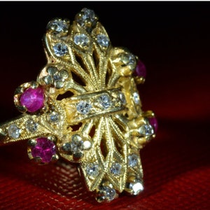 May include: A gold ring with a delicate, intricate design featuring a cross shape. The ring is adorned with small, sparkling diamonds and four ruby stones.