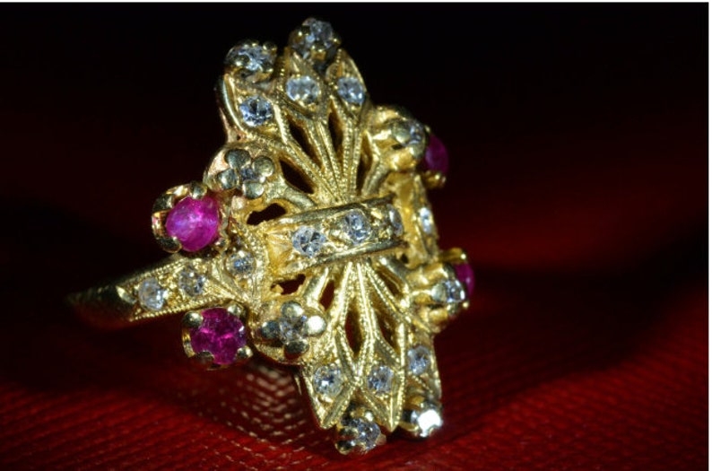 May include: A gold ring with a delicate, intricate design featuring a cross shape. The ring is adorned with small, sparkling diamonds and four ruby stones.