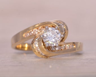 May include: A gold ring with a large, round, clear diamond in the center. The ring is set with smaller diamonds around the center stone and along the band.