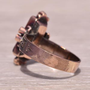 Pu&ograve; includere: Un anello vintage color oro con un gruppo di pietre preziose rosso scuro. La fascia dell'anello ha un design inciso. Le pietre preziose sono incastonate in un'incastonatura decorativa, creando un motivo floreale o a stella. L'anello &egrave; su una superficie chiara.