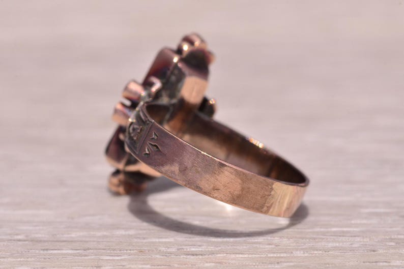 Pu&ograve; includere: Un anello vintage color oro con un gruppo di pietre preziose rosso scuro. La fascia dell'anello ha un design inciso. Le pietre preziose sono incastonate in un'incastonatura decorativa, creando un motivo floreale o a stella. L'anello &egrave; su una superficie chiara.