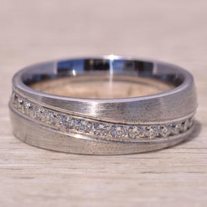 May include: A silver-colored ring with a brushed finish. A row of small, clear gemstones is set diagonally across the ring's band. The ring is on a light-colored wooden surface. Jewelry.