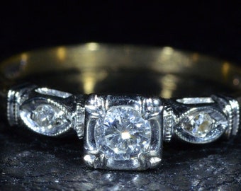 Pode incluir: Anel de noivado vintage com um diamante central de corte quadrado, ladeado por dois diamantes menores em uma cravação ornamentada. A banda é dourada, sobre um fundo escuro.
