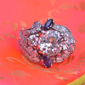 May include: A silver ring featuring a large, round, clear diamond surrounded by smaller diamonds and two blue sapphires. The ring is displayed on a pink and yellow patterned surface.