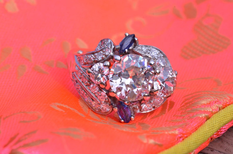 May include: A silver ring featuring a large, round, clear diamond surrounded by smaller diamonds and two blue sapphires. The ring is displayed on a pink and yellow patterned surface.