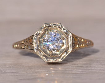 May include: A gold engagement ring with a round diamond set in an octagonal halo. The band is delicate and features intricate filigree details.