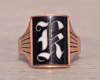 Puede incluir: Anillo cobrizo con una cara negra rectangular con una letra "K" blanca. El anillo tiene un diseño acanalado en los laterales. El anillo está sobre una superficie de madera clara.