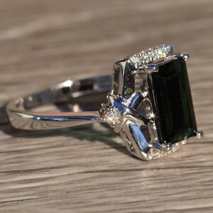 K&ouml;nnte beinhalten: Ein silberner Ring mit einem gro&szlig;en smaragdgr&uuml;nen Edelstein und kleineren klaren Edelsteinen, die in einem filigranen Design gefasst sind.