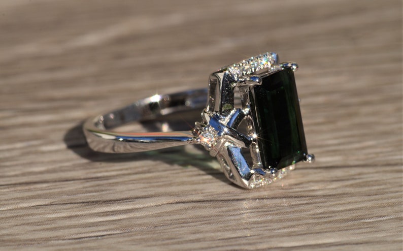 K&ouml;nnte beinhalten: Ein silberner Ring mit einem gro&szlig;en smaragdgr&uuml;nen Edelstein und kleineren klaren Edelsteinen, die in einem filigranen Design gefasst sind.