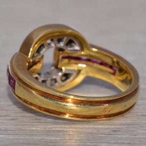 May include: A gold ring with a distinctive design, set with small red gemstones and clear stones in the centre. The ring has a polished finish and a hallmark visible on the band.
