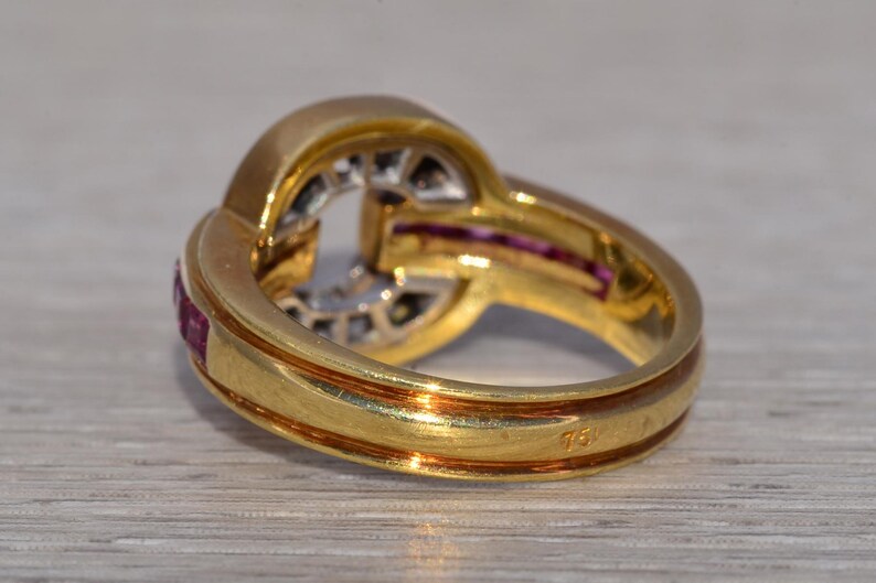 May include: A gold ring with a distinctive design, set with small red gemstones and clear stones in the centre. The ring has a polished finish and a hallmark visible on the band.