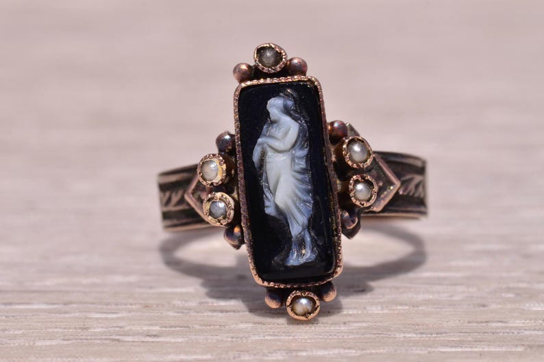 Pu&ograve; includere: Anello antico con cammeo nero rettangolare raffigurante una figura classica bianca. L'anello ha una montatura color oro con piccole perle e accenti decorativi. La fascia ha un design inciso.