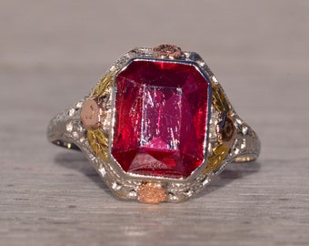 May include: A vintage ring with a large, deep red gemstone set in a filigree design. The ring is made of white gold with accents of yellow and rose gold.