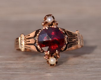 May include: A gold ring with a red gemstone and two white pearls. The ring has a detailed design with intricate patterns.