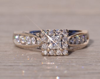 May include: A silver engagement ring with a square-shaped diamond center stone surrounded by smaller diamonds. The band is set with smaller diamonds.