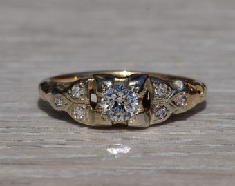 Pode incluir: Um anel de ouro com um diamante central redondo e transparente. O anel tem um design decorativo com diamantes menores e detalhes florais na banda. O anel está sobre uma superfície de madeira clara.