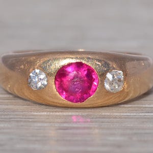 May include: A gold ring with a large, round, bright pink center stone and two smaller, clear stones on either side. The ring has a wide band and a brushed finish, set against a light wood grain surface.