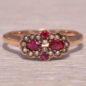 Peut inclure: Une bague dorée ornée d'un groupe de pierres rouges et de petites perles blanches. Le motif central de la bague comprend deux rubis ovales, entourés de petites pierres et perles, sur une surface claire.