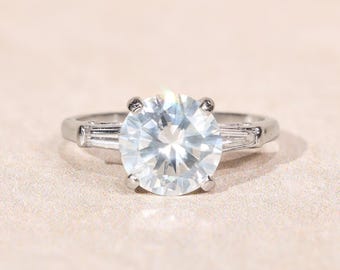 May include: A silver-toned ring featuring a large, round, brilliant-cut clear center stone held by four prongs. The band is accented on each side by a single, elongated, tapered baguette-cut clear stone, creating a classic three-stone design.