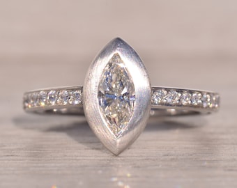 Puede incluir: Un anillo de plata con un diamante de forma marquesa engastado en un engaste de bisel. El anillo tiene una banda de diamantes más pequeños.