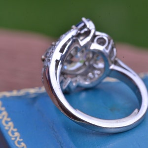 May include: A silver ring featuring a large, clear centre stone surrounded by smaller stones. The ring is displayed in a blue velvet box.