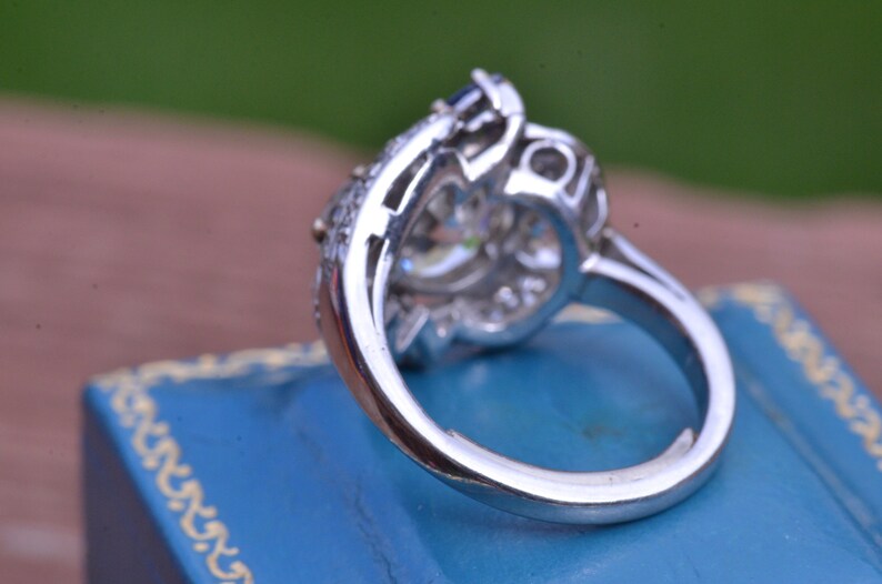 May include: A silver ring featuring a large, clear centre stone surrounded by smaller stones. The ring is displayed in a blue velvet box.