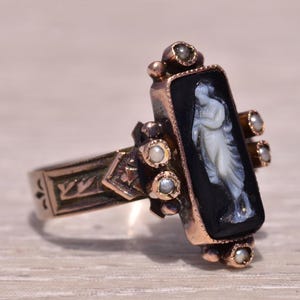 Pu&ograve; includere: Un anello d'oro vintage con un cammeo nero rettangolare raffigurante una figura bianca. L'anello &egrave; impreziosito da piccole perle e dettagli intricati sulla fascia. Il design dell'anello &egrave; classico ed elegante.