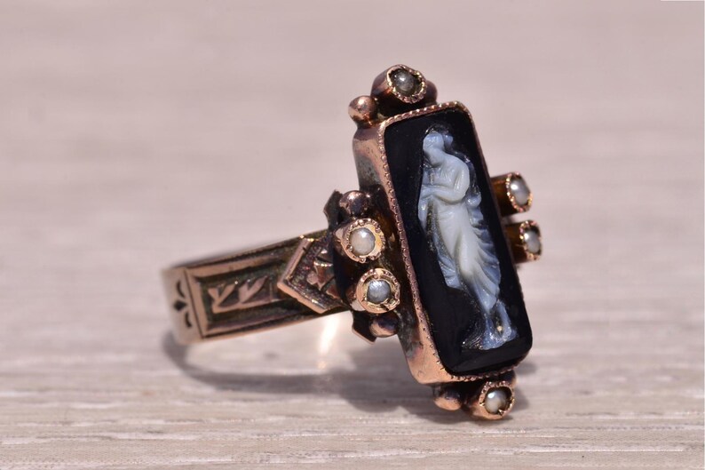 Pu&ograve; includere: Un anello d'oro vintage con un cammeo nero rettangolare raffigurante una figura bianca. L'anello &egrave; impreziosito da piccole perle e dettagli intricati sulla fascia. Il design dell'anello &egrave; classico ed elegante.