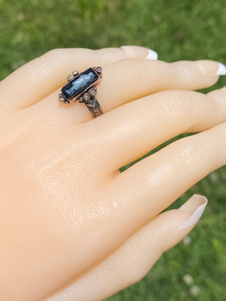Pu&ograve; includere: Un anello vintage in argento con una pietra rettangolare blu scuro con un design cameo bianco. L'anello &egrave; adornato con piccole perle rotonde e dettagli decorativi attorno alla pietra e sulla fascia. L'anello &egrave; esposto su una mano con unghie curate.