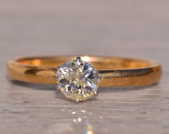 Puede incluir: Un anillo de oro con un gran diamante transparente en el centro. El diamante es de talla redonda y está engastado con cuatro garras. La banda es de oro pulido simple. El anillo se muestra sobre una superficie clara.
