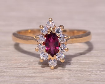 Op de afbeelding: Een gouden ring met een marquise-geslepen robijn als middensteen, omringd door een halo van ronde, heldere edelstenen. De ring heeft een eenvoudige, gepolijste gouden band. De edelstenen zijn in een bloemachtig patroon geplaatst.