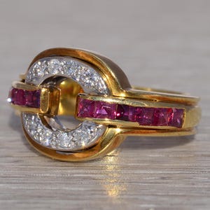 May include: A gold ring with a distinctive design. The ring has a central, circular feature with a band of small, square-cut rubies and a band of small diamonds. The ring's band is also gold.