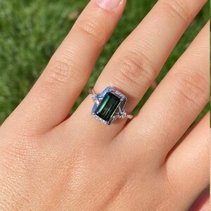 K&ouml;nnte beinhalten: Ein silberner Ring mit einem gr&uuml;nen Smaragd-Schliff-Edelstein, der in einem zarten, verzierten Rahmen gefasst ist. Der Ring ist mit kleineren Diamanten um den Edelstein und den Rahmen verziert.
