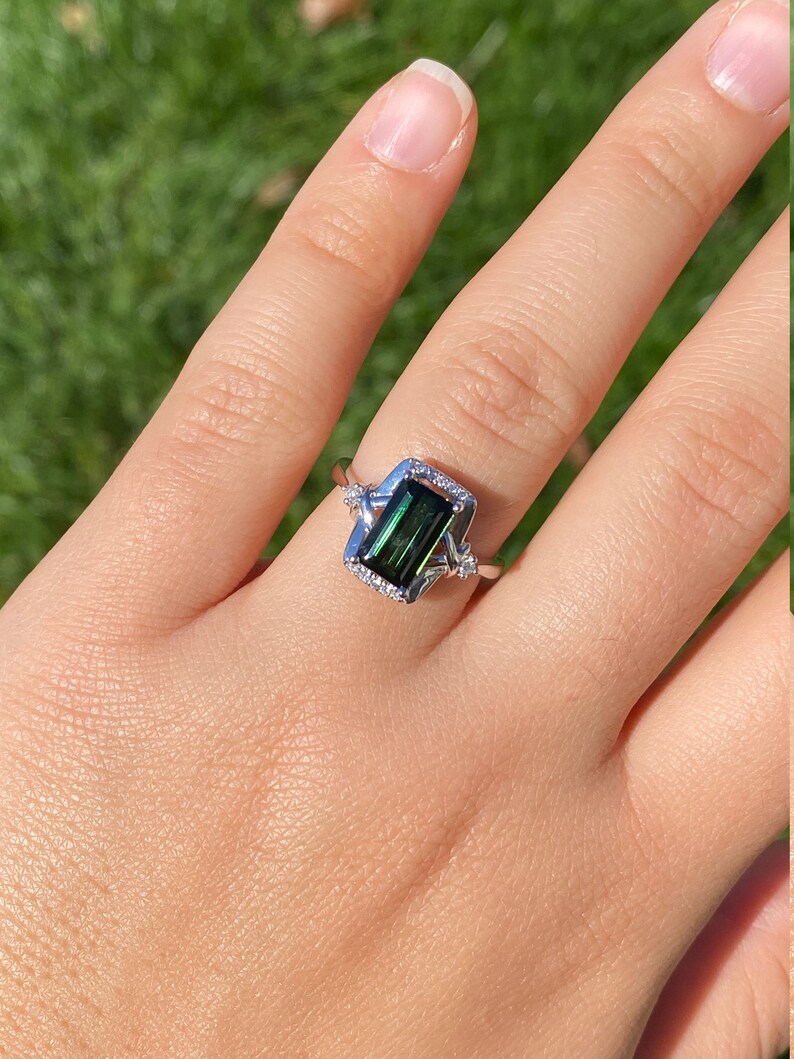 K&ouml;nnte beinhalten: Ein silberner Ring mit einem gr&uuml;nen Smaragd-Schliff-Edelstein, der in einem zarten, verzierten Rahmen gefasst ist. Der Ring ist mit kleineren Diamanten um den Edelstein und den Rahmen verziert.