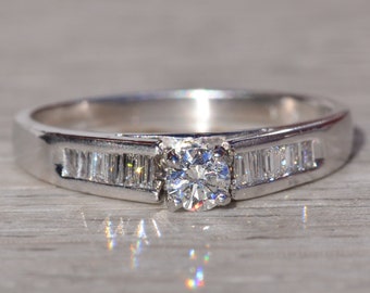 May include: A silver engagement ring with a round center diamond and a band of smaller diamonds set in a channel setting.